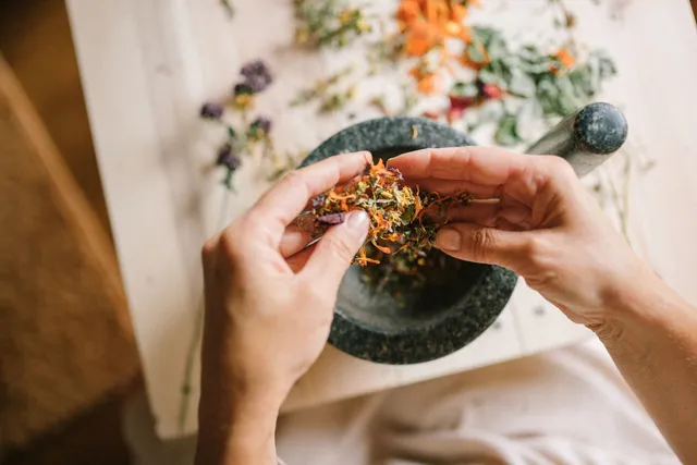 Hands crumbling dried herbs into a stone mortar on a wooden table