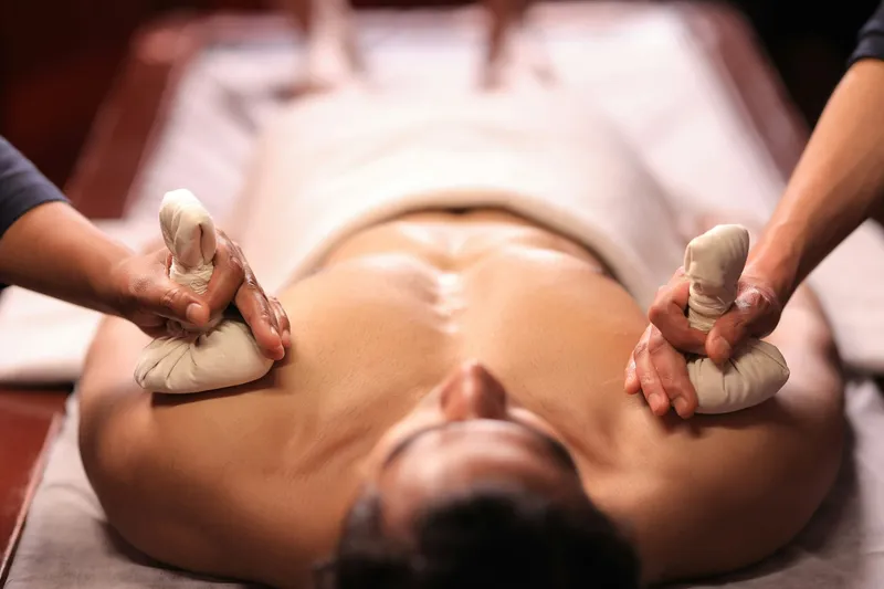Therapist pressing two warm herbal poultice bundles onto an oiled back on a wooden treatment table