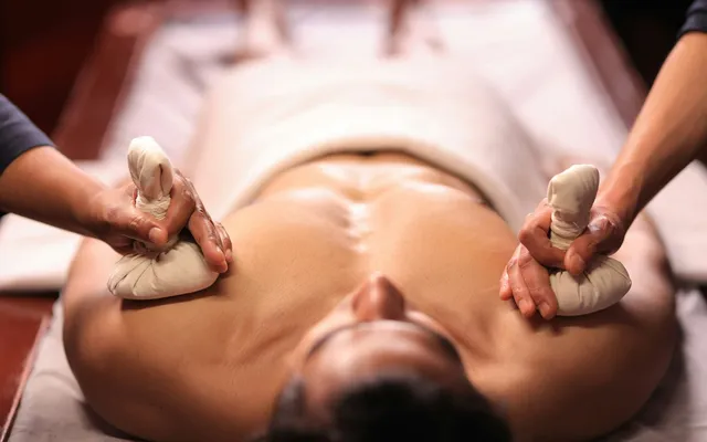 Therapist pressing two warm herbal poultice bundles onto an oiled back on a wooden treatment table