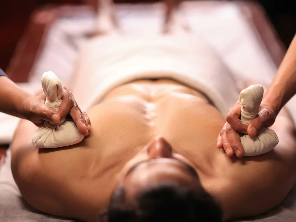 Therapist pressing two warm herbal poultice bundles onto an oiled back on a wooden treatment table