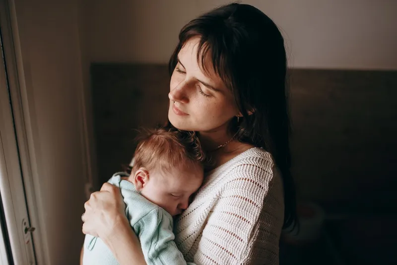 A mother holding a sleeping newborn by a window in soft daylight