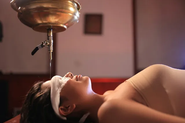 Shirodhara: a thin stream of warm oil poured from a brass vessel onto the forehead of a patient whose eyes are covered
